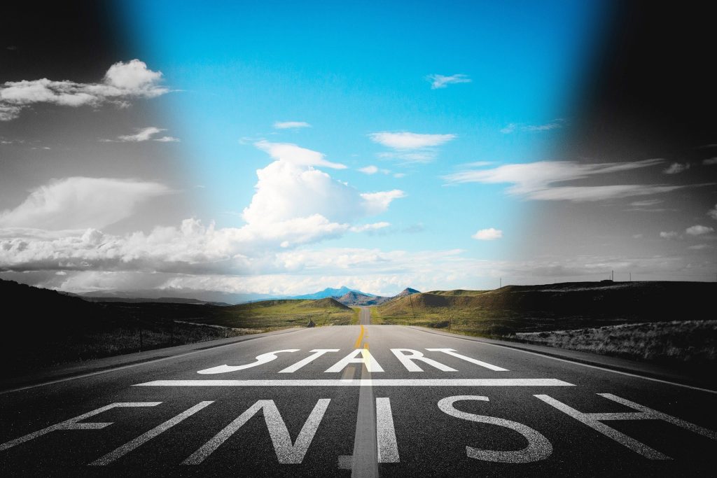 A road leading towards some mountains. The sky in the centre is blue and around the edges black and white. Start / Finish is written in white paint on the road.