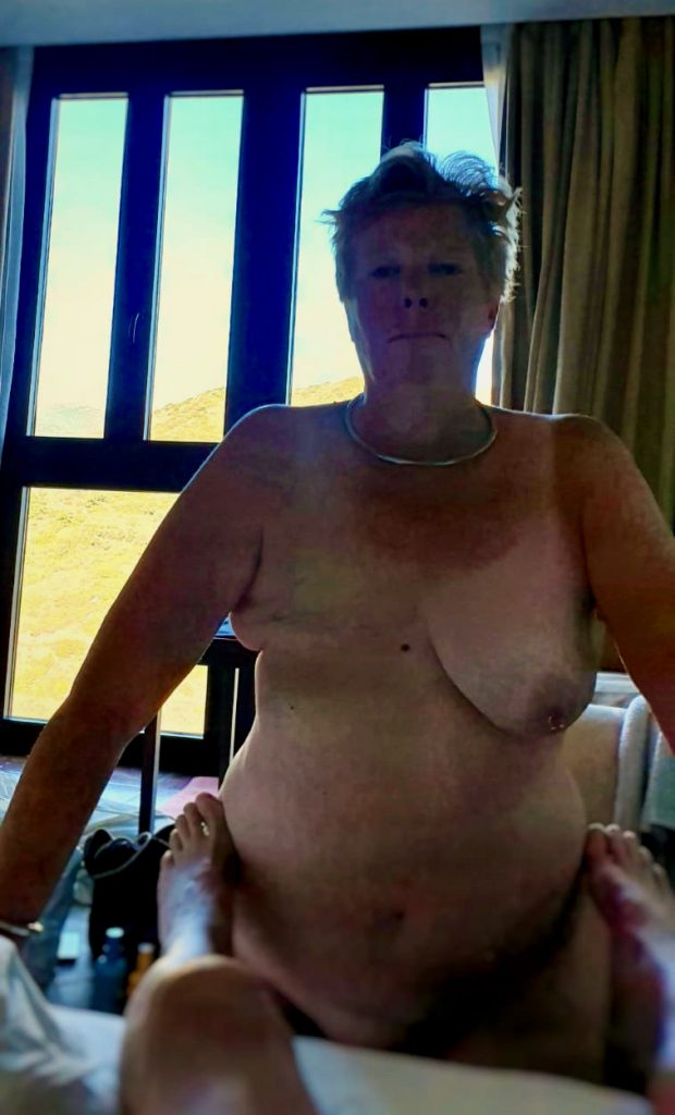 I am standing naked. I don't look happy, but I might have just woken up and been tired. I'm standing with my hips facing the bed and Master's feet are between my hips. It's a hotel room, but I don't know where or when it was taken. 
