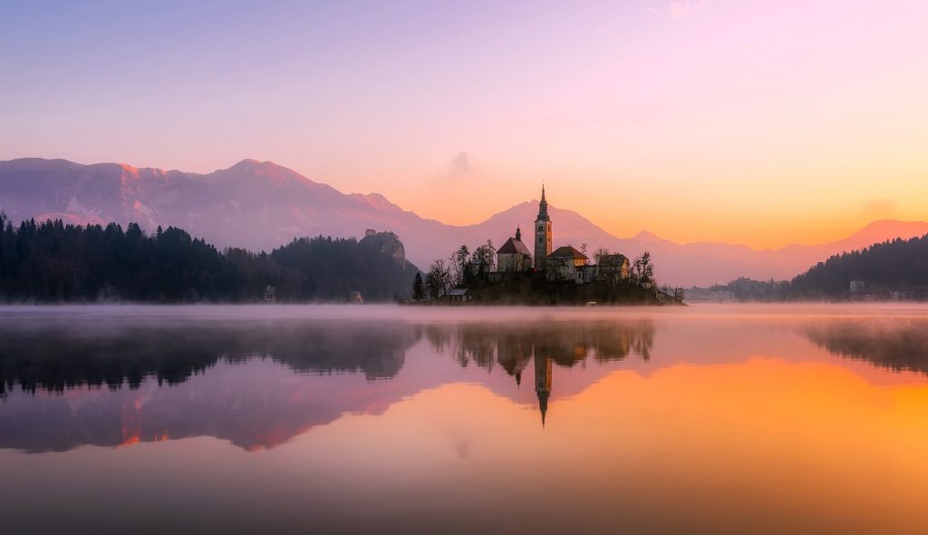 body of water near mountain during daytime with a church at the centre. The image is reflected into the water 