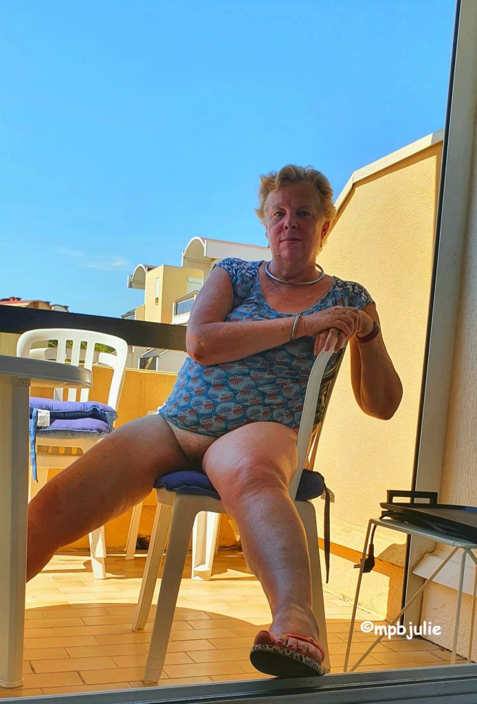 Me sitting on a chair on the balcony of an apartment in France. It is a hot sunny day, with blue sky. I am wearing a blue and red top, but no underwear. My legs are spread. 
