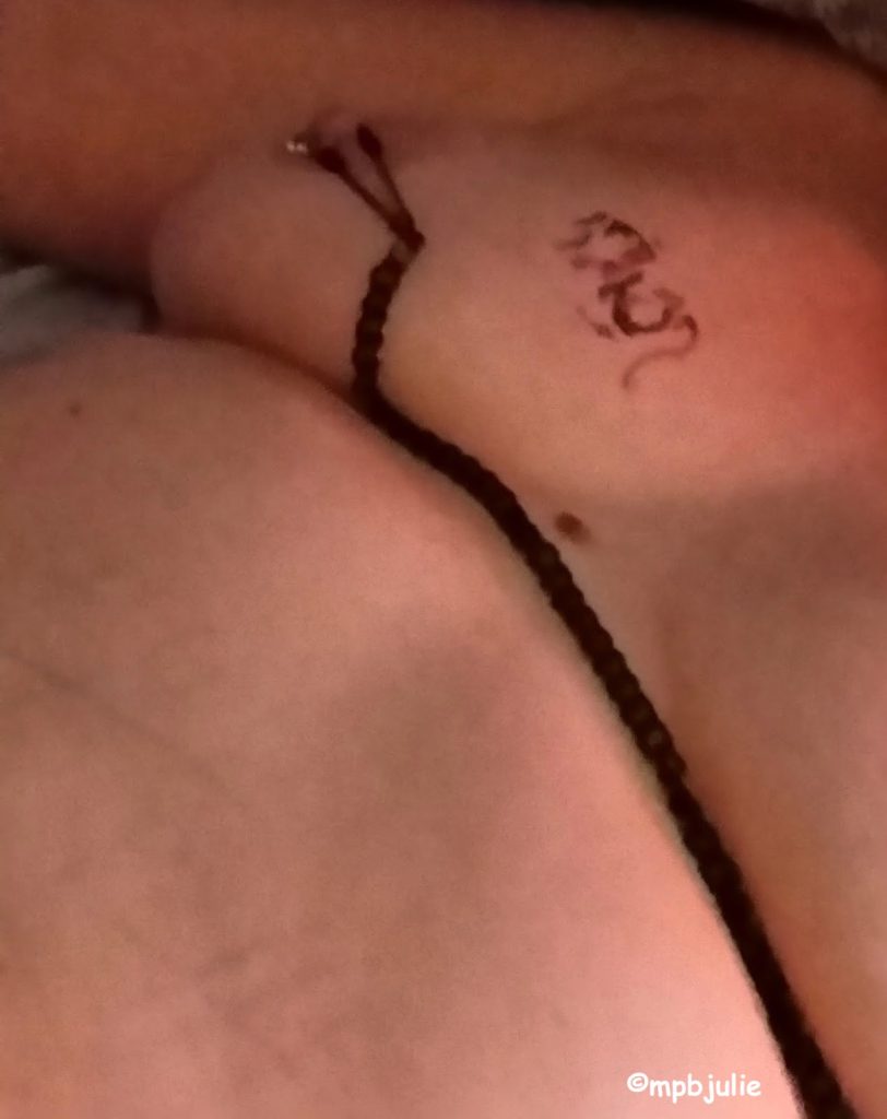 I'm lying on my back. You can see one breast, with nipple piercing. Some rope is entwined around the nipple and runs across the breasts. There is a faded tally mark on my tummy and a faded tattoo on the breast. 