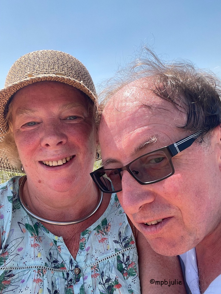 This is a selfie of us. I'm wearing a hat, a smile, my collar and a green flowery top. He is looking at the camera in a cool way, slightly sideways on. Glasses dark and a half smile. 