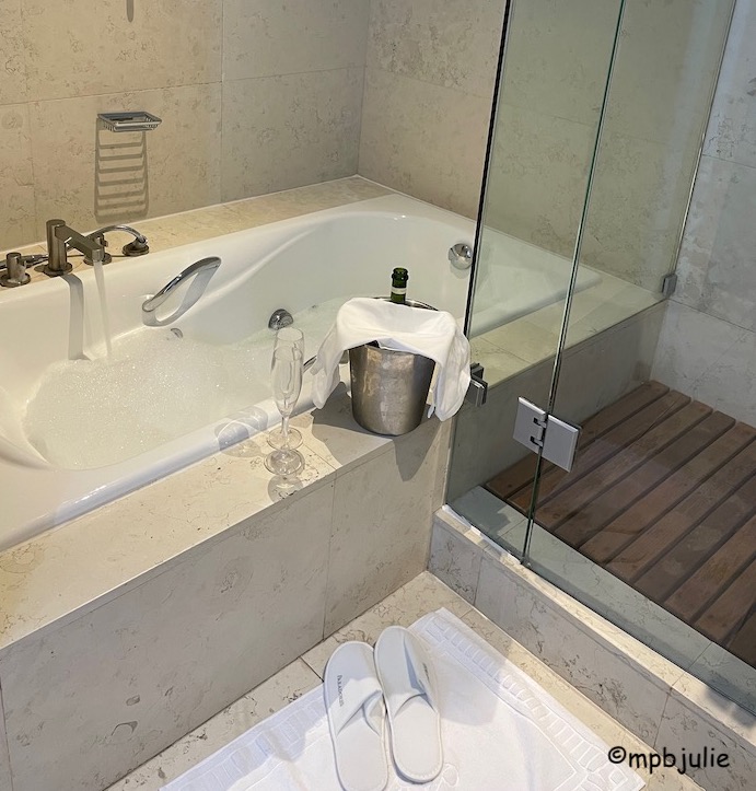 The image shows a bath filling with water and a few bubbles forming. On the side of the bath is a wine bucker containing a bottle of cava, covered with a towel. There are two glasses (plastic). On the floor is a white mat and some parador slippers. To the right you can see there is a separate shower in this bathroom.