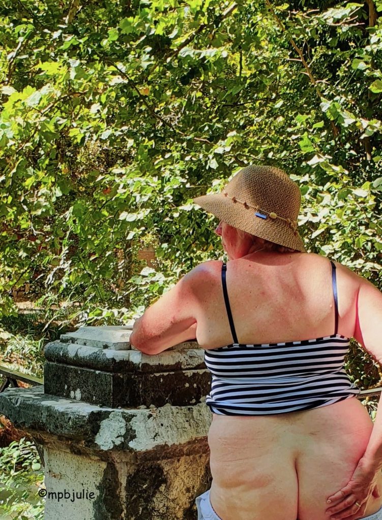 I'm standing leaning on a weathered stone post with greenery from trees in the background. I'm wearing a sun hat and blue and white stripy sun top, pulled up to my waist. My bottom is bare and my hand rests on my right butt cheek. I'm facing the bushes. 