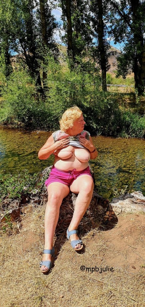 Me seated on a large log or tree stump Wearing pink shorts and blue sandals. My top is pulled up so you can see my boobs.