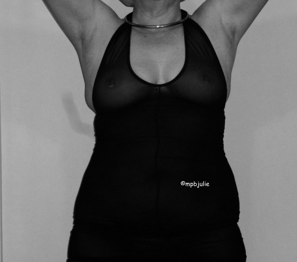 A black and white image of Mme standing with arms raised. I'm wearing a figure hugging see through. black dress. My collar is visible but not may face. My right nipple piercing can be seen and cleavage.