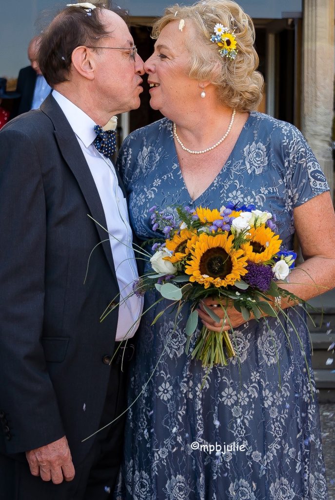 Wedding photo. We are poised to kiss. Heads covered in confetti. He is wearing a dark suit and bow tie, white shirt. I'm wearing a blue and white flowery dress and carrying a bouquet of sunflowers.