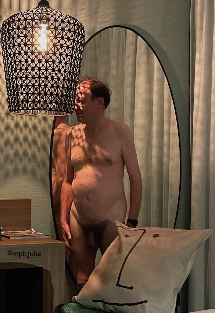 A hotel room shot of a naked Master. H is looking directly at a lamp shade that is in the shape of a large up side down basket. He is by a mirror that reflects his arm and shoulder. 