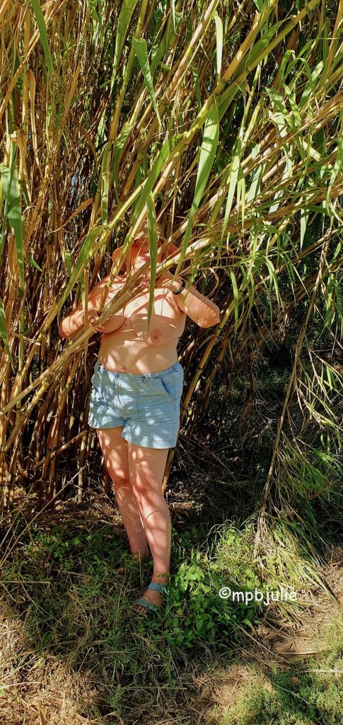 I'm standing partially hidden by. some bamboo. I'm topless, but wearing. a straw hat, denim shorts and sandals. 