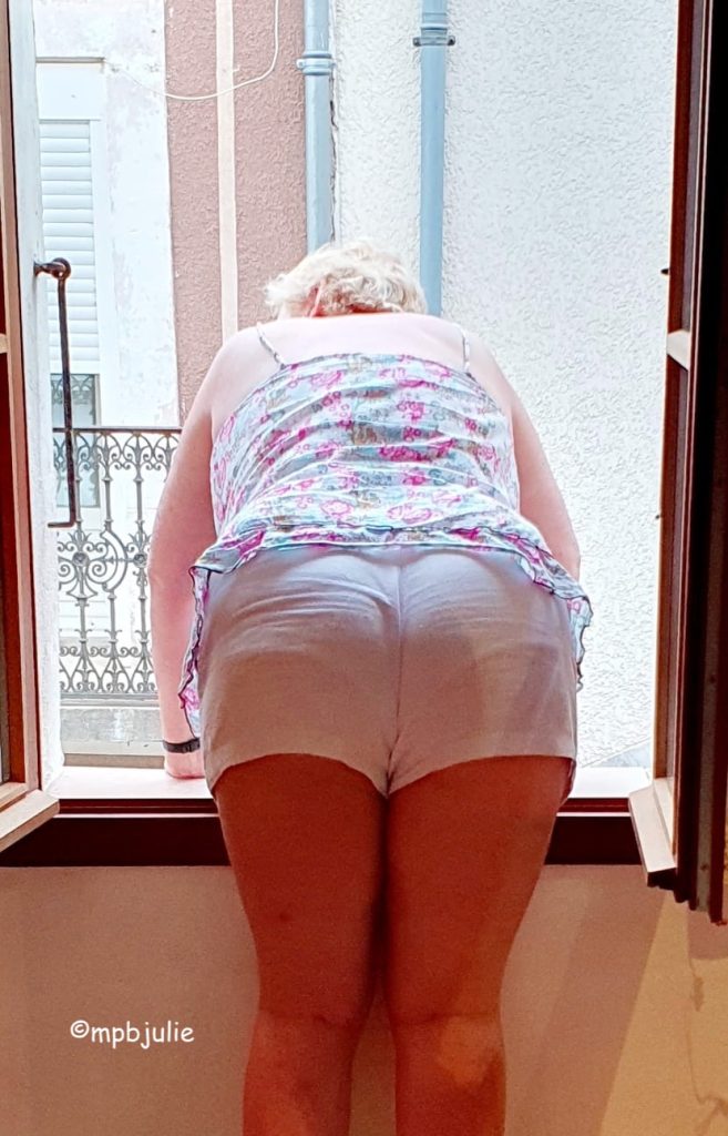 I'm fully dressed in this image, but wearing white shorts. and a strappy top. I'm leaning out of the. kitchen window of our house in France to the street beyond.