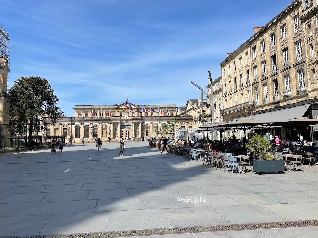Large square in Bordeaux. Buildings to right and at the back. Right hand ones have bars and restaurants and tables outside with people sitting in some. 