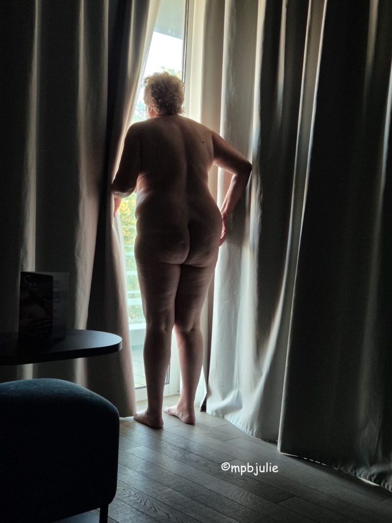 I'm looking out of a full length window. The curtains are still closed on two sides so there's just a small gap for me and a bit of. light. I'm naked
