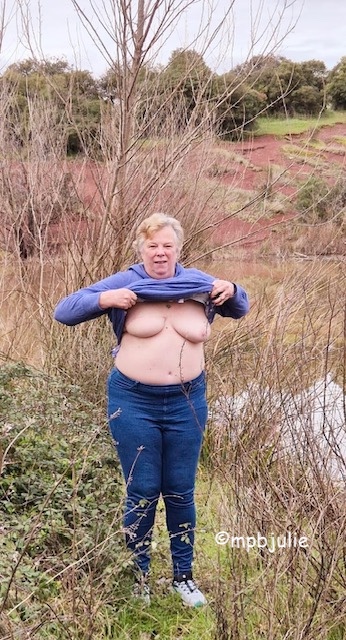 I'm standing in amongst some brushwood and behind me is a brown looking lake. In the background trees and some clay soil. I'm flashing my tits while wearing a purple hoody and blue jeans. 