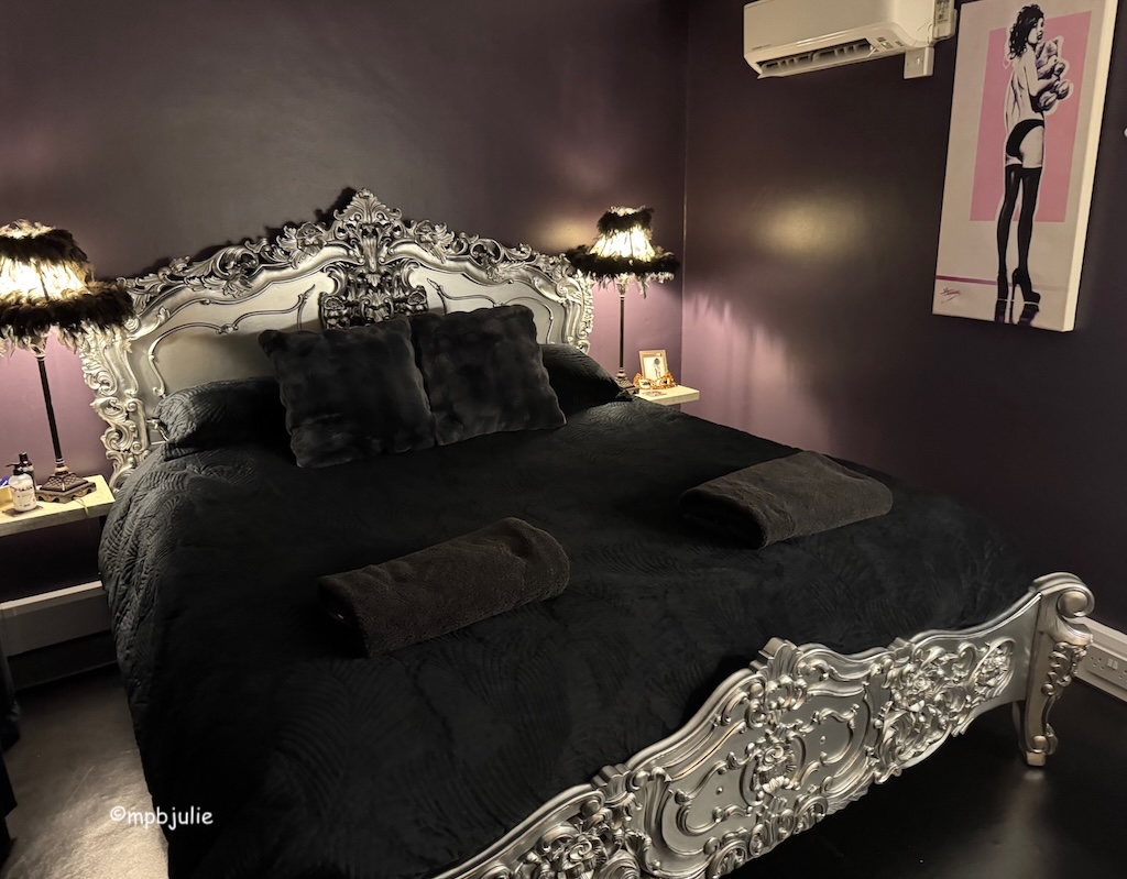 A silver bed frame with black furry cushions and bed cover. Black / purple walls and bedside lamps frilled with black lace. 
